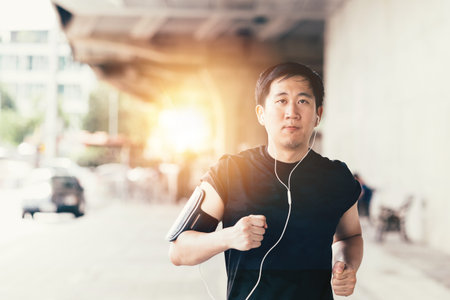 Young determined athletic Asian man wearing headphone and armband with smart phone inside. He is...