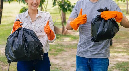 Cropped young happy Asian male and female couple volunteers with gloves giving thumbs up while collecting garbage waste into trash bag in green park. Environmental friendly and litter collectionの写真素材