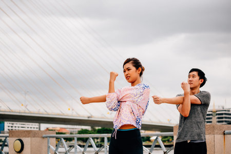 Asian man and woman stretching arms and looking away while standing on bridge against overcast sky...