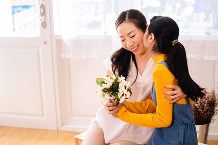 Face of Asian teenage daughter hugging and kissing happy smiling middle-aged mother with tenderness in indoor living room at home. Mother is holding a bouquet received from child. Mothers day conceptの写真素材
