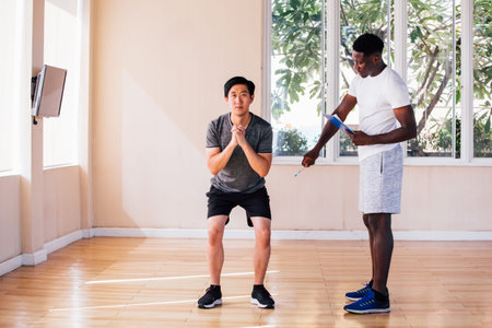 Asian young sportsman doing squat exercise while African American personal trainer watching in gym...