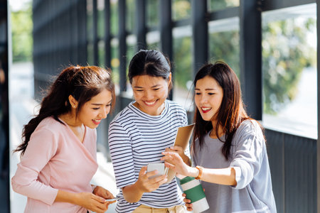 Young Asian business women talking and showing phone screen in office building in casual wear. Three girls having discussion outdoorsの写真素材