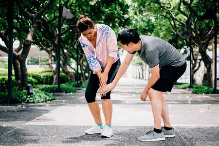 Asian male touching aching knee of woman in sportswear while helping during accident on training in parkの写真素材