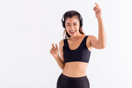 Happy Asian woman in black sport bra listening to music and dancing with raised hands on white...