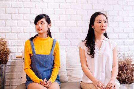Unhappy mother and daughter in bad relationship sitting together and looking aside on brick wall backgroundの写真素材