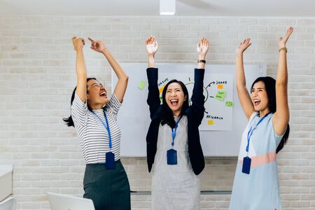 Smiling Asian young and mature business women standing in line with arms raised up gesture in meeting room with excited feeling. Row of Business people portraitの写真素材