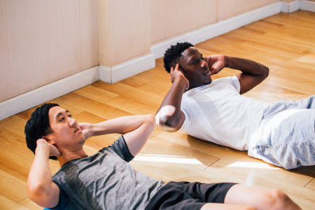 From above multiethnic athletic men lying on floor holding hands behind head doing and abdominal...