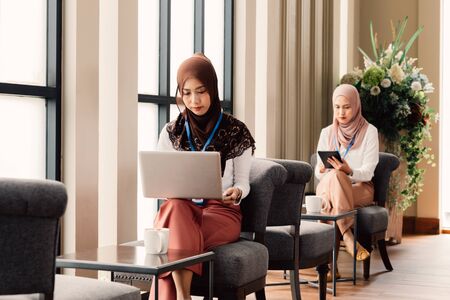 Islamic Southeast Asian ladies in hijabs using laptop and tablet while doing job in stylish elegant restaurantの写真素材