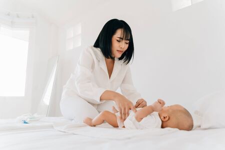 Young Asian mother with closed eyes in white wear changing diaper for own infant lying on bed in houseの写真素材