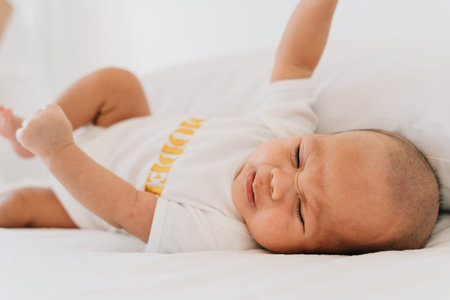 Sad small Asian infant in pajama with closed eyes crying newborn baby lying on bed at home