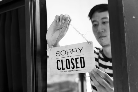 Through glass view of Asian male business owner hanging up sign with inscription closed at end of...