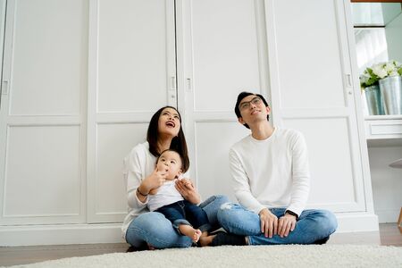 From below of positive young Asian married couple with baby boy looking up admiringly while sitting together on floor against white wardrobe in roomの写真素材
