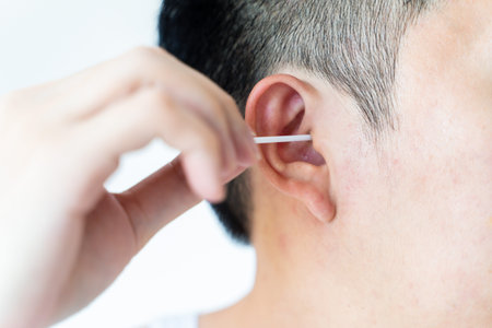 Closeup of black haired man washing ears with cotton bud in toilet on white background