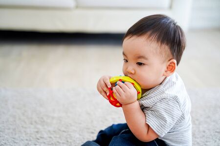 Side view of adorable serious calm Asian toddler boy in striped shirt and jeans sucking colorful toy while sitting on carpet at homeの写真素材