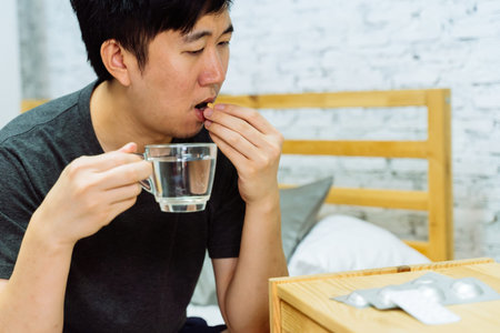 Young Asian man feeling sick and taking pills while sitting on the bed at home with medicines tablets and drinking water on table. Healthcare and Medicine conceptの写真素材