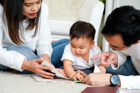 Happy modern Asian parents sitting on floor with cute infant boy and reading fairy tale while...