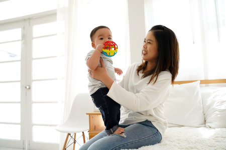 Asian family of mother playing toy doll with their son on bed at home. Young woman carrying baby boy...