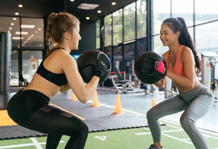 Happy young female athletic people performing squat exercises with friend and holding a medicine ball at fitness gym. Group of two confident women with healthy lifestyle working out togetherの写真素材