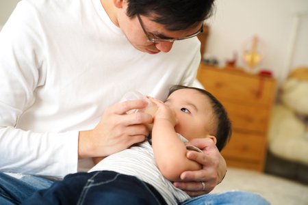 Asian family of young father feeding a baby boy from milk bottle at home