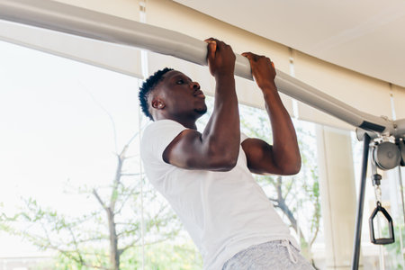 Strong African American sportsman in white T-shirt doing pull ups during fitness workout in modern...