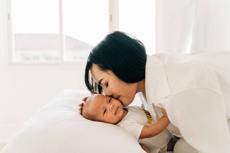Side view of caring young Asian mum in white wear holding own infant kissing newborn son at homeの写真素材