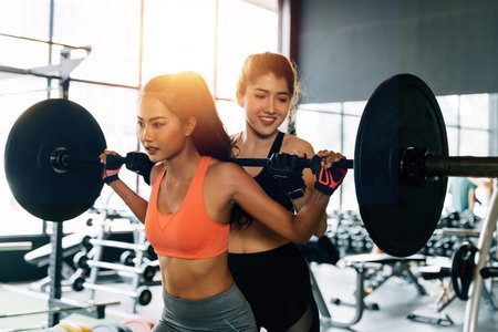Asian personal trainer coaching a bodybuilding woman to perform the exercise squat with barbell in the fitness gym. Weightlifting and workout training conceptの写真素材