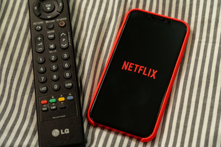 BANGKOK, THAILAND - MARCH 27, 2020: Man sitting on bed in bedroom holds Netflix logo on smart phone...
