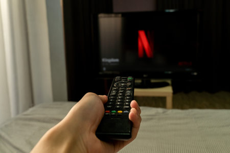 BANGKOK, THAILAND - MARCH 27, 2020: Man sitting on bed in bedroom holds Netflix logo on smart phone screen. Netflix is an entertainment company, provides streaming media and video on demand online.のeditorial素材