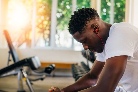 Side view of tired African American man in sportswear listening to music in wireless earphones while sitting on bench and resting during training in gym.の写真素材