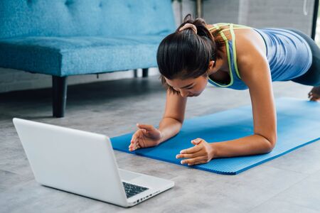 20s young Asian woman in sportswear doing plank poses while watching fitness training class on computer laptop online. Healthy girl exercising and learning in living room. Internet education concept.の写真素材