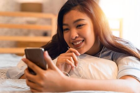 Portrait of happy young Asian girl in casual clothing lying down on bed while making a video call with smartphone and waving in bedroom at home. Video conferencing technology concept.の写真素材