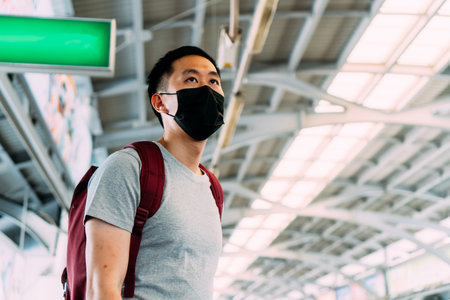 Close up of one young Asian man wearing a black surgical face mask waiting for the train during new...