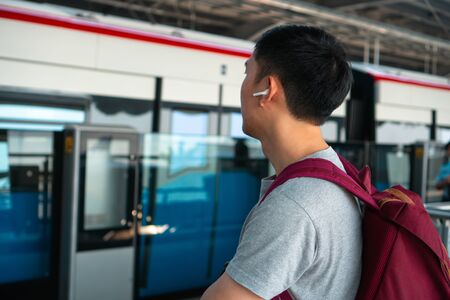 Close up of young man listening to music with wireless earpods while commuting by train at station. Asian guy enjoying music on the go.の写真素材