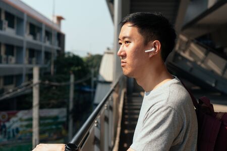 Close up of young man listening to music with wireless earpods while commuting by train at station. Asian guy enjoying music on the go.の写真素材
