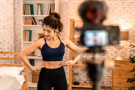 20s young Asian woman in sportswear doing talking to camera and explaining fitness tutorials online. Healthy girl blogger in living room at home. Teaching online bodybuilding class concept.の写真素材