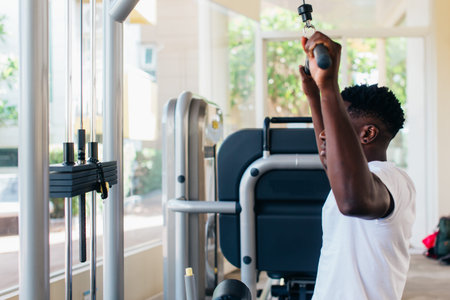 Strong African American man in sportswear pulling cable with bar while sitting on exercise machine...