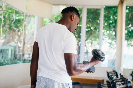Young African American man standing and lifting a dumbbell with the rack at gym. Male weight...