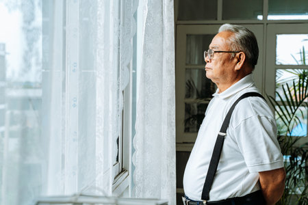 Close up of pensive old man with glasses looking out of window feeling lonely. 70s Asian elderly...