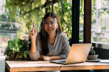 Young 20s Asian woman looking at camera while wearing a protective mask with laptop, mobile phone, and notebook for working outside in casual business look. Corona Virus - Covid 19 work online conceptの写真素材
