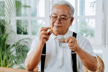 Senior Asian man with cup of water and pill taking remedy in cozy room at home. Old male taking medicineの写真素材