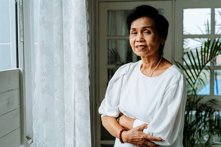 Positive elderly ethnic female with folded arms smiling and looking at camera while standing near...