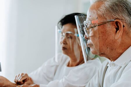 Side view of elderly Asian man and woman in face shields resting in light room during coronavirus pandemicの写真素材