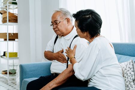 Senior woman taking care of elderly Asian man suffering from heartburn while sitting on sofa in cozy living room at home. Old pensioner sick patientの写真素材
