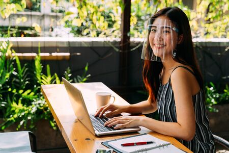Young Asian woman looking at camera while wearing a protective mask with laptop, mobile phone, and notebook for working outside in casual business look. Corona Virus - Covid 19 work online concept.の写真素材