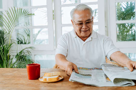 Happy senior Asian male in casual clothes and eyeglasses reading interesting article in newspaper while having breakfast at home. Elderly man enjoying retirement lifestyle.の写真素材