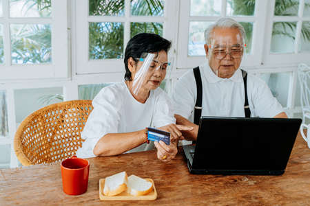 Elderly Asian couple in personal protective face shield sitting at table at home. Senior man and woman with PPE using laptop and credit card while making online shopping during coronavirus pandemic.の写真素材