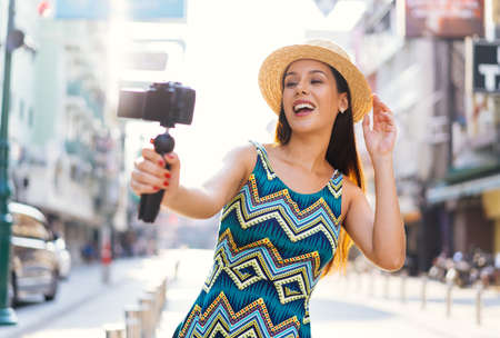 Young brunette woman using a video camera to record journey with smile. Happy and excited 20s Hispanic girl traveler wear summer dress with hat waving to internet audience. Influencer vlogging conceptの写真素材
