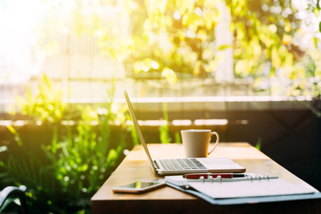 Modern laptop and smartphone placed on table near open planner with pen and cup of hot beverage on sunny summer day on terraceの写真素材