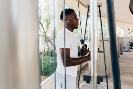 Muscular African American sportsman doing cable fly with exercise machine standing against window...
