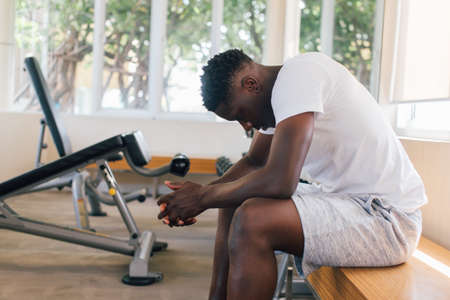 Desperate African American male athlete sitting on bench during break in fitness training in modern gymの写真素材